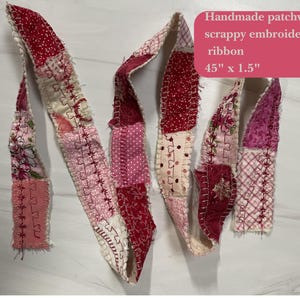 May include: Handmade patchwork scrappy embroidered ribbon in shades of red, pink, and cream. The ribbon is 45 inches long and 1.5 inches wide. The ribbon is made of various fabric scraps and features decorative stitching.