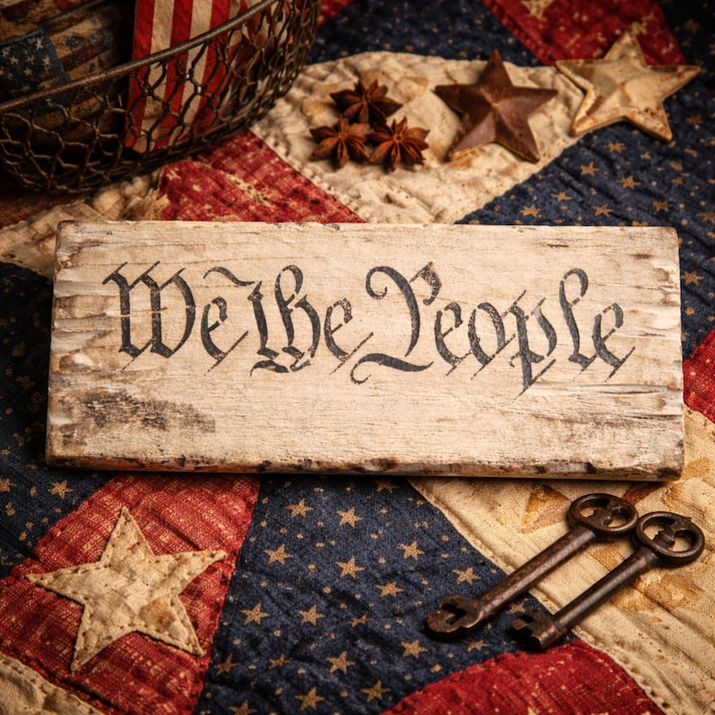 May include: A rustic wooden sign with the words "We the People" in black script. The sign is weathered and sits on a patriotic quilt with star and stripe patterns. Two antique keys are placed below the sign.