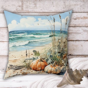 May include: A decorative throw pillow featuring a beach scene with two pumpkins on the sand. The ocean is blue with white waves and a light blue sky with white clouds. The pumpkins are orange and have green stems.