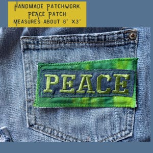 May include: A handmade patchwork denim patch with the word "PEACE" embroidered in green thread. The patch measures approximately 6 inches by 3 inches.