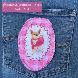 May include: A pink and white fabric patch with a chihuahua wearing a pink sweater. The patch is oval shaped and has a stitched border. The text "HANDMADE BRUISER PATCH 4.25" x 3" is printed on a pink banner above the patch.