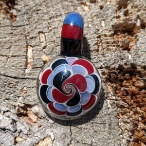 May include: A colorful glass pendant with a spiral design. The pendant features a blue, red, and black top, transitioning to a round body with a spiral of red, black, and white petals. The pendant is set against a textured wooden surface.
