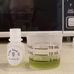 May include: A small translucent plastic bottle labeled Grape Seed Oil by Raws 2 Pindures Plus next to a clear plastic measuring cup containing a light green liquid. The cup features black measurement markings at 5 mL, 10 mL, and 15 mL increments.