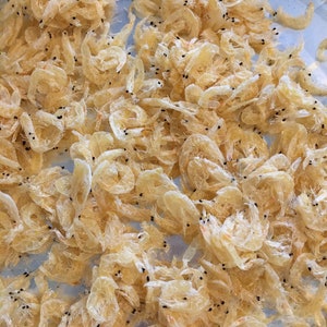 May include: A close-up of a pile of dried, pale yellow shrimp. The shrimp are arranged in a random pattern and are all about the same size.