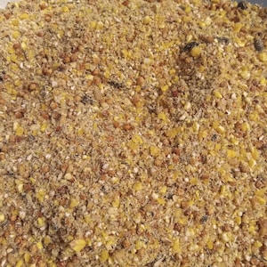 May include: A coarse, dry mixture of animal feed consisting of small, granular particles in shades of tan, light brown, and bright yellow. The texture is uneven and varied, featuring a blend of crushed grains, seeds, and small organic fragments.