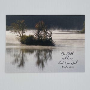 May include: A serene landscape print featuring a small island with trees reflected in calm water. The image has a soft, misty quality with the text "Be Still and know that I am God Psalm 46:10" in the lower right corner.