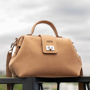 May include: A tan leather handbag with a top handle and a detachable shoulder strap. The bag features a rectangular shape with a flap closure and silver-colored hardware. The initials "JMB" are embossed on the flap.