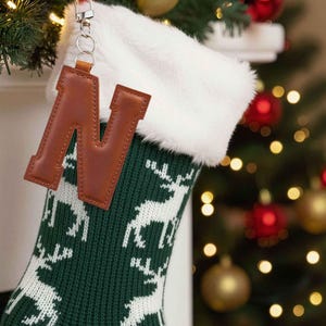 May include: A green Christmas stocking with a white reindeer pattern and a faux fur cuff. A brown leather letter "N" charm hangs from the stocking. The stocking is hanging near a decorated Christmas tree.