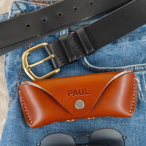 May include: A brown leather sunglasses case with the name "PAUL" embossed on it, a pair of black round sunglasses, and a black leather belt with a gold buckle, all resting on blue denim jeans.