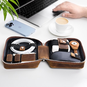 May include: A brown leather travel case with a zipper closure, open to reveal a variety of compartments and accessories, including a coiled cable, a small pouch, and a charging cable.