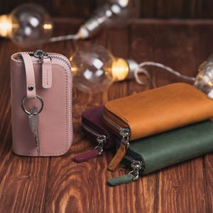 May include: Leather key wallets in shades of pink, brown, and green, each with a silver zip. One pink wallet is open, revealing keys. The wallets are displayed on a wooden surface, with fairy lights in the background.