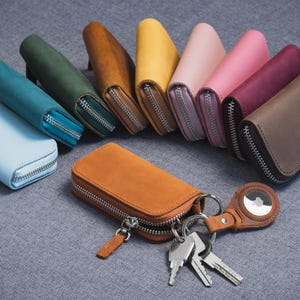 May include: A collection of colorful leather key cases with silver zippers. The cases are in various colors, including blue, green, brown, yellow, pink, and burgundy. A tan key case with keys and a leather AirTag holder is in the foreground.