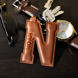 May include: A brown leather keychain shaped like the letter "N", with "NOAH" embossed. It's next to a watch, keys, and sunglasses on a dark wooden surface. The keychain is a personalised accessory.