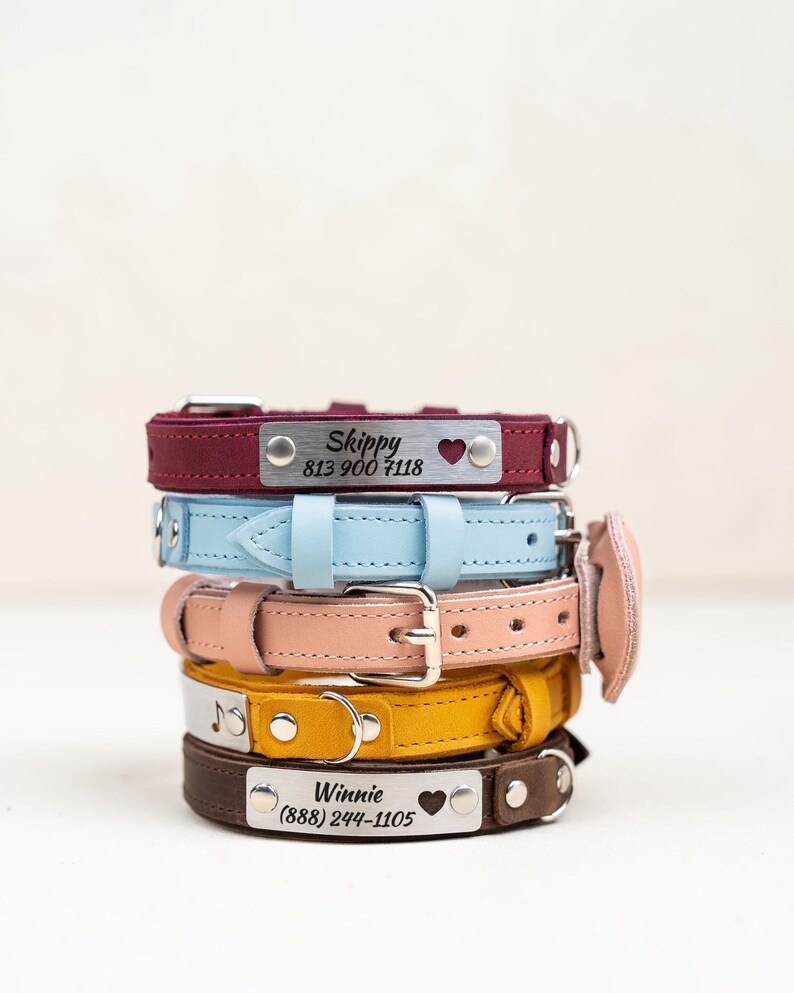 Op de afbeelding: Een stapel gepersonaliseerde hondenhalsbanden in verschillende kleuren: bordeauxrood, lichtblauw, perzik, geel en bruin. Elke halsband heeft een zilveren naamplaatje met een naam, telefoonnummer en hartsymbool. De halsbanden zijn van leer.