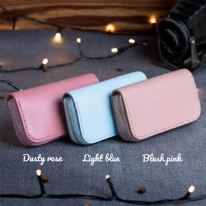 May include: Three leather zip-around wallets in dusty rose, light blue, and blush pink. Each wallet has a silver zipper and a rectangular shape. The wallets are displayed on a gray surface with string lights.