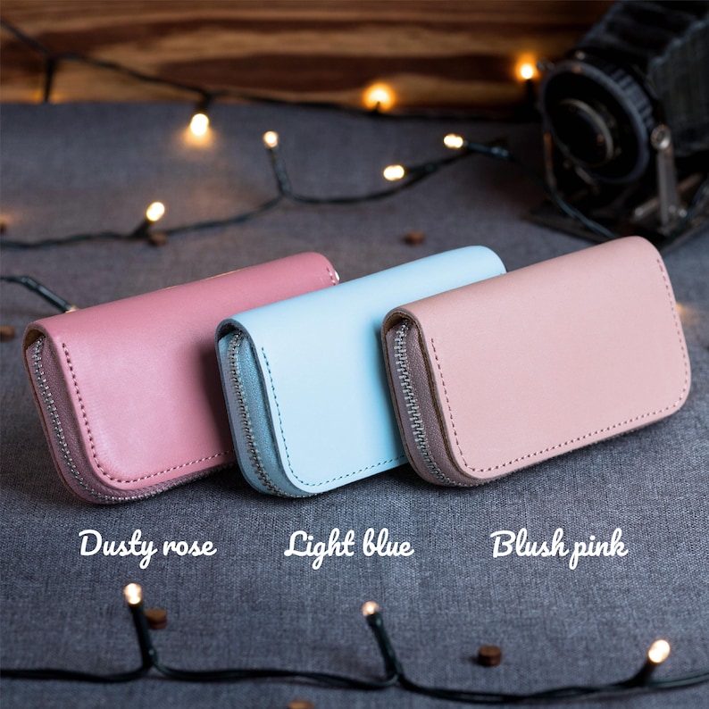 May include: Three leather zip-around wallets in dusty rose, light blue, and blush pink. Each wallet has a silver zipper and a rectangular shape. The wallets are displayed on a gray surface with string lights.