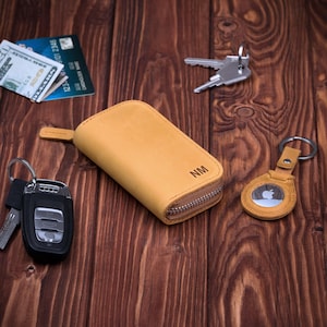 May include: A tan leather key case with a zipper closure and the initials "NM" is displayed on a wooden surface. A matching keychain with an Apple AirTag is next to the key case. Other keys, credit cards, and cash are also visible.