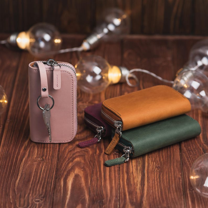 May include: A collection of leather key wallets in various colors, including pink, brown, and green, with silver zippers. One pink wallet is open, displaying keys. The wallets are arranged on a wooden surface, with string lights in the background.