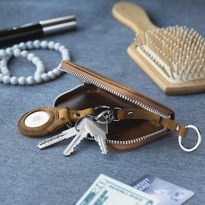 May include: A brown leather key case with a silver zipper, holding keys and an Apple AirTag. A white beaded bracelet, a wooden hairbrush, and a pen are also visible on a gray surface.