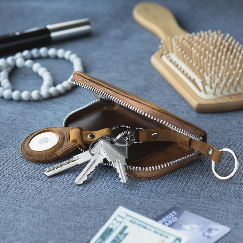 May include: A brown leather key case with a silver zipper, holding keys and an Apple AirTag. A white beaded bracelet, a wooden hairbrush, and a pen are also visible on a gray surface.
