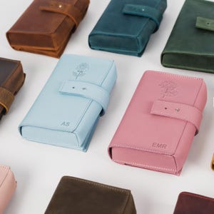 May include: Assortment of leather-bound journals in various colors, including brown, blue, green, pink, and light blue. Each journal features a floral design and a personalized monogram. The journals are arranged on a white surface.