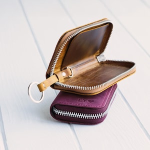 May include: Two leather key wallets, one open and one closed, are stacked on a white surface. The open wallet is tan with a silver zipper and key ring. The closed wallet is a deep purple with a silver zipper and the letters "LY" and a heart symbol.