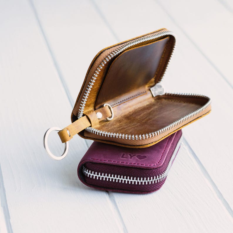 May include: Two leather key wallets, one open and one closed, are stacked on a white surface. The open wallet is tan with a silver zipper and key ring. The closed wallet is a deep purple with a silver zipper and the letters "LY" and a heart symbol.