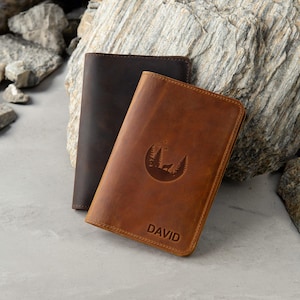 May include: Two leather passport holders, one brown and one dark brown. The brown passport holder has a moon and trees design with the name "DAVID" embossed on the front.