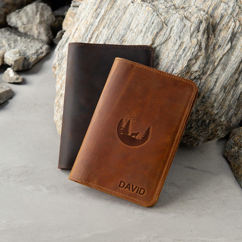 May include: Two leather passport holders, one brown and one dark brown. The brown passport holder has a moon and trees design with the name "DAVID" embossed on the front.