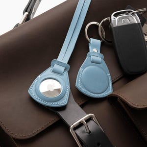 May include: A brown leather briefcase with a blue leather strap and two blue leather keychains. One keychain has a clear circle with an Apple logo. The other keychain has a solid blue circle. A black leather belt with a silver buckle is attached to the briefcase.