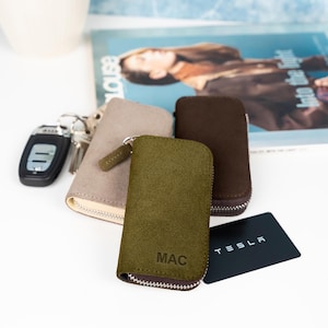 May include: A collection of keychains and key cases in various colors. The key cases are olive green, brown, and tan, with a zipper closure. The olive green case has the initials "MAC" embossed on it. A black Tesla card is also visible.