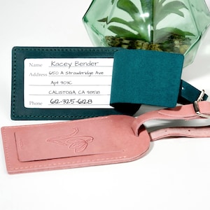 May include: A teal leather luggage tag with a white label that says "Kacey Bender" and includes an address and phone number. The tag is attached to a pink leather luggage tag with a floral design.