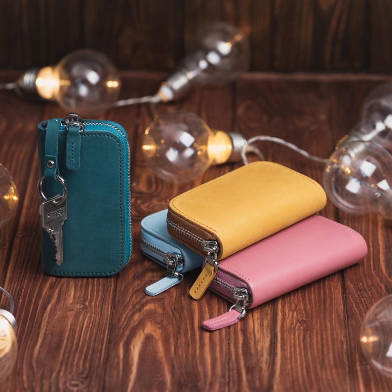 May include: A collection of colorful leather key wallets in teal, light blue, yellow, and pink, each with a zipper closure. One teal wallet has a key attached. The wallets are arranged on a wooden surface, with decorative string lights in the background.