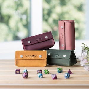 May include: A collection of personalized leather keychains in various colors: burgundy, pink, green, and yellow. Each keychain is rectangular with a snap closure and features a name: "LEO", "EMMA", "LUCK", and "TOM". Dice are scattered around the keychains.