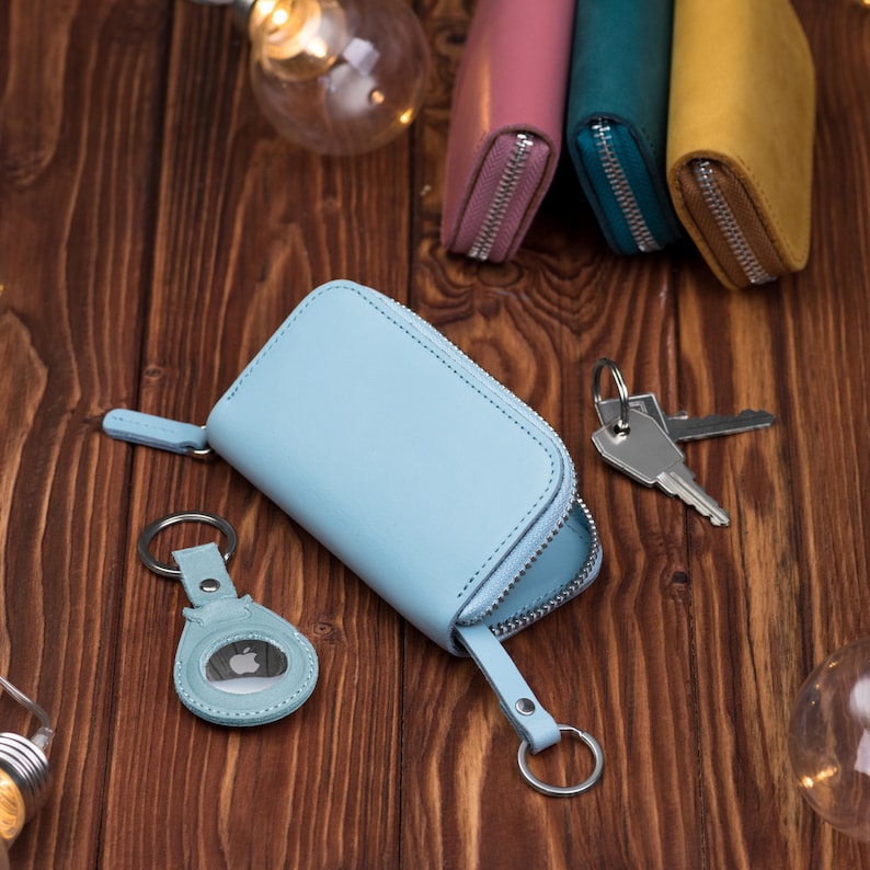 May include: A light blue leather key case with a silver zipper and a matching key fob. The key case is open, revealing the interior. Other key cases in pink, teal, and yellow are visible in the background.