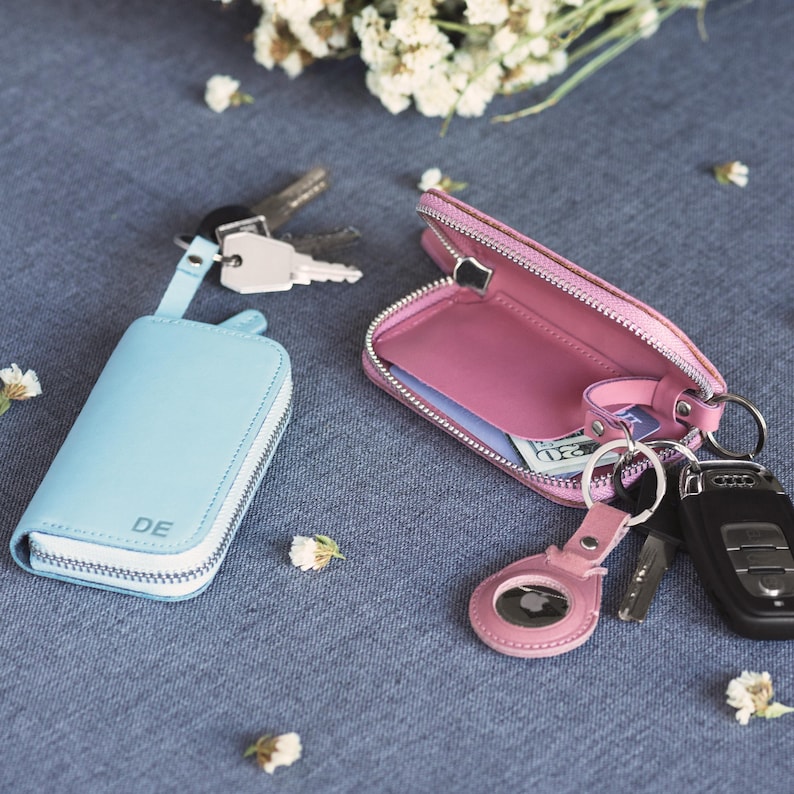 May include: Two leather key cases, one light blue and one pink, with silver zippers. The light blue case has the letters "DE" on it. Keys and a key fob are attached to the cases. The pink case is open, revealing a small amount of cash.