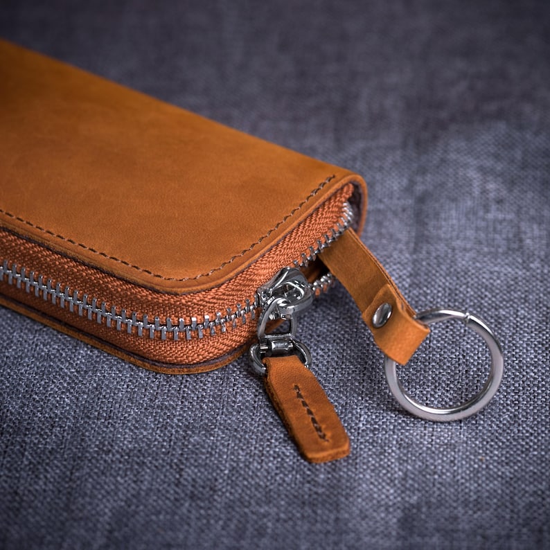 May include: Close-up of a brown leather wallet with a silver zipper and key ring. The wallet is rectangular and has a stitched border. The zipper pull and key ring loop are also made of brown leather.