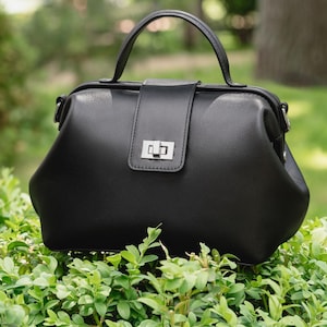May include: A black structured handbag with a smooth finish, featuring a single top handle, a rectangular front flap, and a silver-toned metal turn-lock closure. The bag has a wide, rounded base and a frame-style opening at the top.