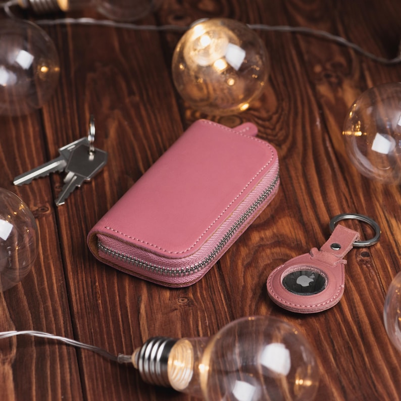 May include: A pink leather key case with a silver zipper, a matching AirTag holder, and a set of keys. The items are on a wooden surface with string lights and decorative light bulbs.