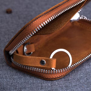 May include: Open brown leather key case with a silver zipper and key ring. The interior is a lighter shade of brown. The case is partially open, revealing the key ring and interior details. The zipper is silver.