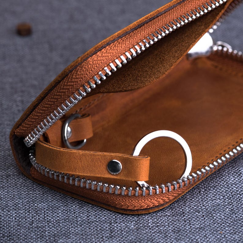 May include: Open brown leather key case with a silver zipper and key ring. The interior is a lighter shade of brown. The case is partially open, revealing the key ring and interior details. The zipper is silver.