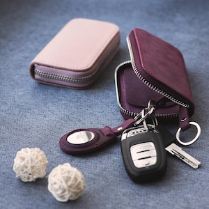 May include: Two leather key cases, one light pink and one deep purple, with silver zippers. The purple case is open, displaying keys and a key fob. Two small, white decorative balls and a purple key ring are also present.