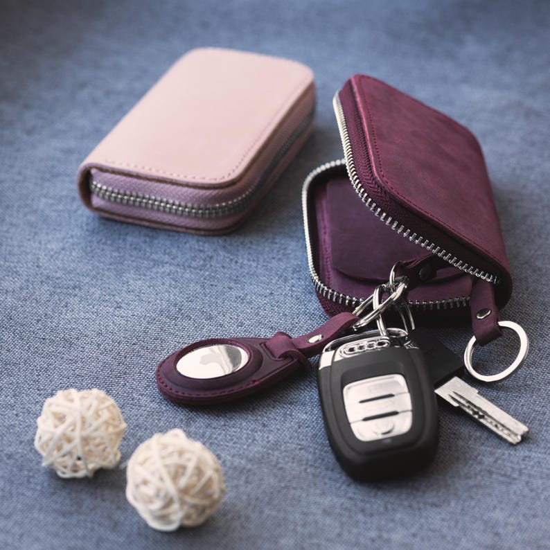 May include: Two leather key cases, one light pink and one deep purple, with silver zippers. The purple case is open, displaying keys and a key fob. Two small, white decorative balls and a purple key ring are also present.