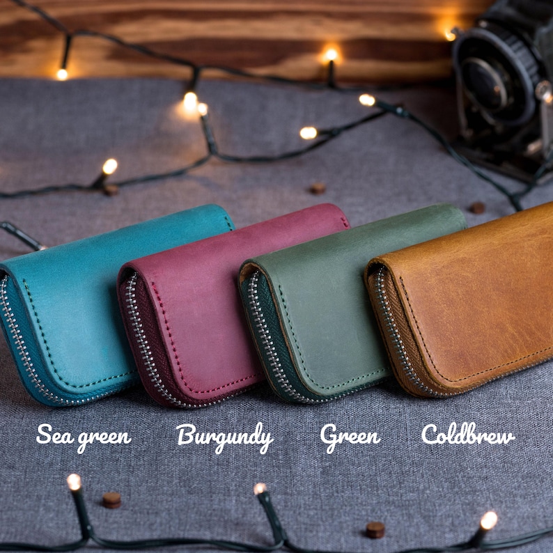 May include: Four leather zip-around wallets in sea green, burgundy, green, and coldbrew colors. Each wallet has a silver zipper and matching stitching. The wallets are displayed on a gray surface.