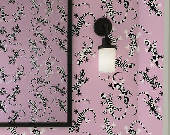 May include: A bathroom with a light pink wall featuring a repeating pattern of black and white lizards. A black framed mirror hangs above a white vanity with a black faucet and sconces. A white towel hangs on a black towel ring.