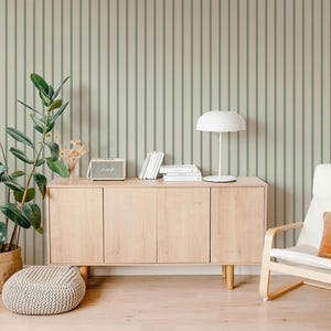 May include: A modern living room scene with a light wood cabinet, a white chair with an orange pillow, and a round knitted ottoman. A potted plant and a white lamp add to the decor. The wall has a vertical striped pattern.