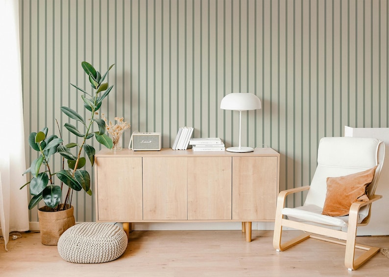 May include: A modern living room scene with a light wood cabinet, a white chair with an orange pillow, and a round knitted ottoman. A potted plant and a white lamp add to the decor. The wall has a vertical striped pattern.