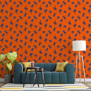 Op de afbeelding: Oranje behang met een herhalend patroon van blauwe kikkerillustraties. Het behang bevindt zich achter een blauwgroene bank met gele kussens, een kleine zwarte tafel en een plant in een pot. Rechts staat een witte lamp met een houten standaard.