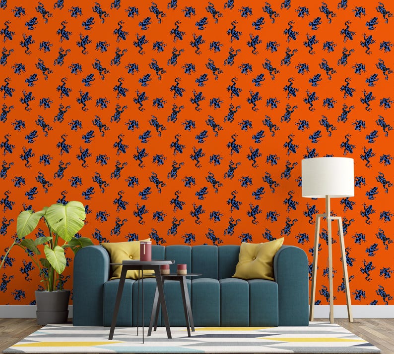 Op de afbeelding: Oranje behang met een herhalend patroon van blauwe kikkerillustraties. Het behang bevindt zich achter een blauwgroene bank met gele kussens, een kleine zwarte tafel en een plant in een pot. Rechts staat een witte lamp met een houten standaard.