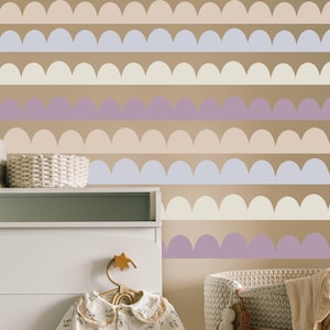 Puede incluir: Una pared decorada con filas horizontales de formas festoneadas en tonos beige, lavanda y azul claro. Una cómoda blanca, una cesta de mimbre y un conjunto de bebé en una percha de madera están en primer plano.